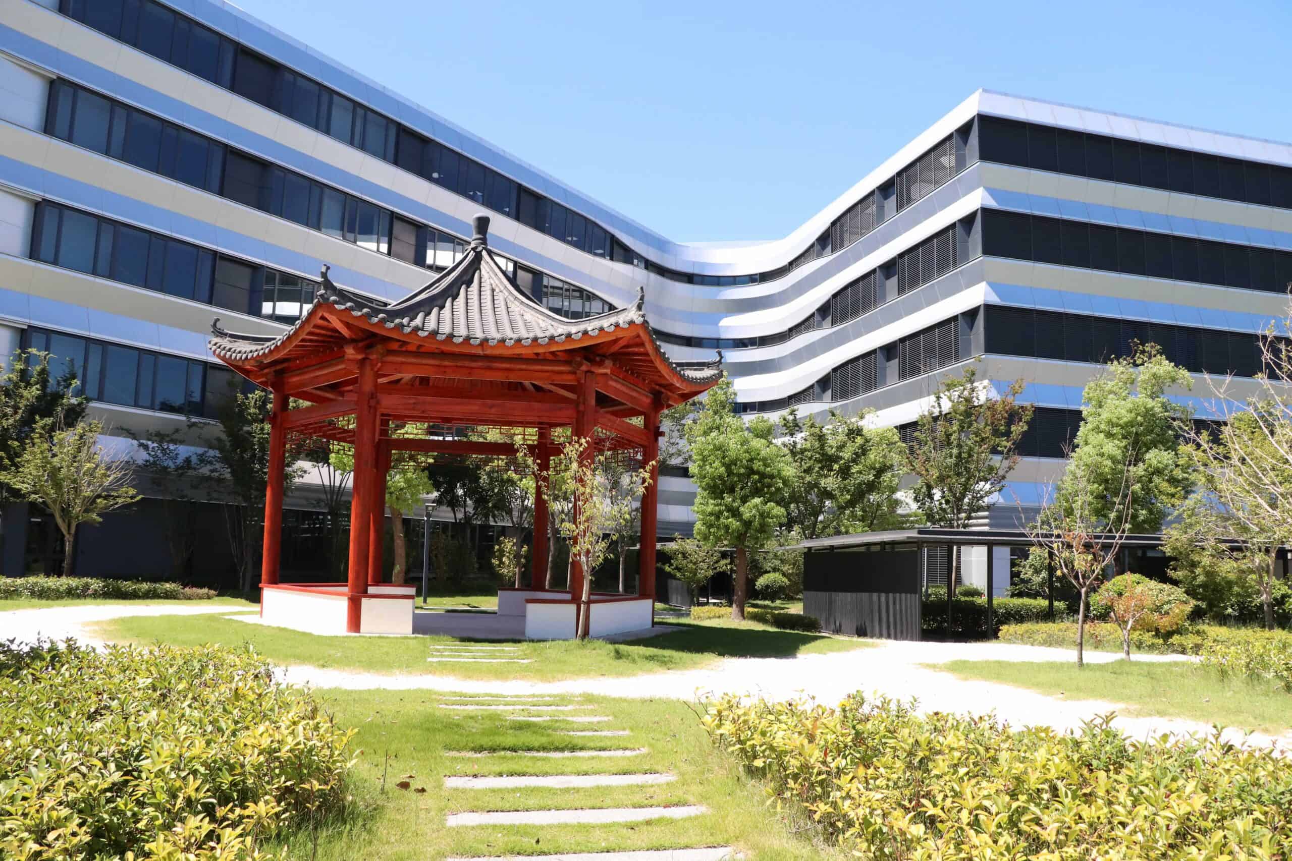 Modern corporate building with traditional Asian garden pavilion in front, green landscaping, bright sunlight.