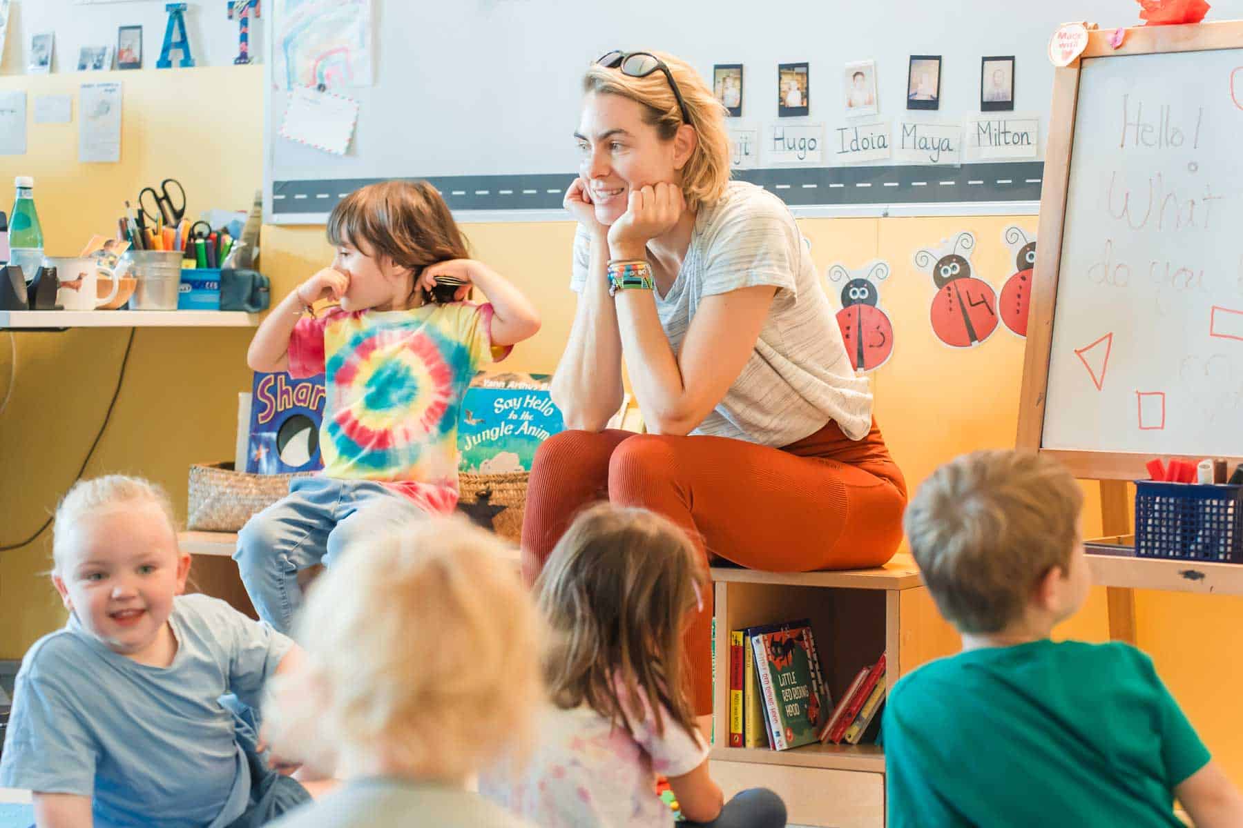 Friendly teacher interacting with diverse young students in a colorful classroom environment.