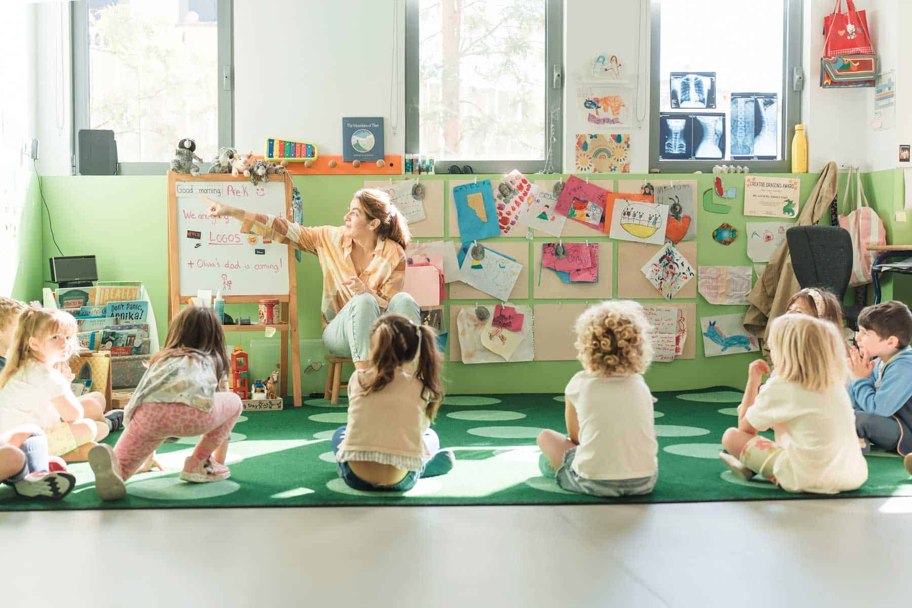 Bright preschool classroom with children sitting on a green rug listening to teacher's storytelling on diverse topics.