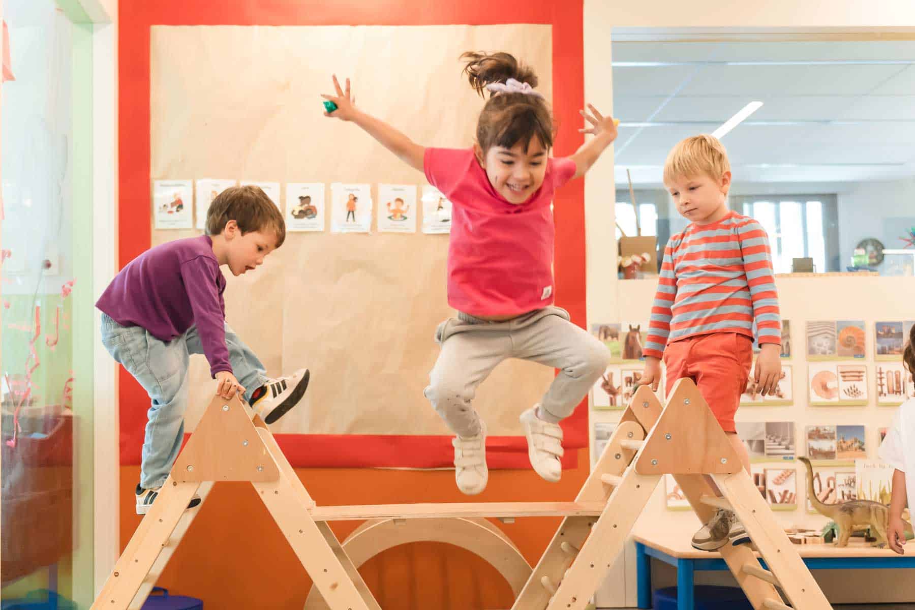 Playful children jumping on a wooden activity structure in a vibrant, modern classroom at a world school.