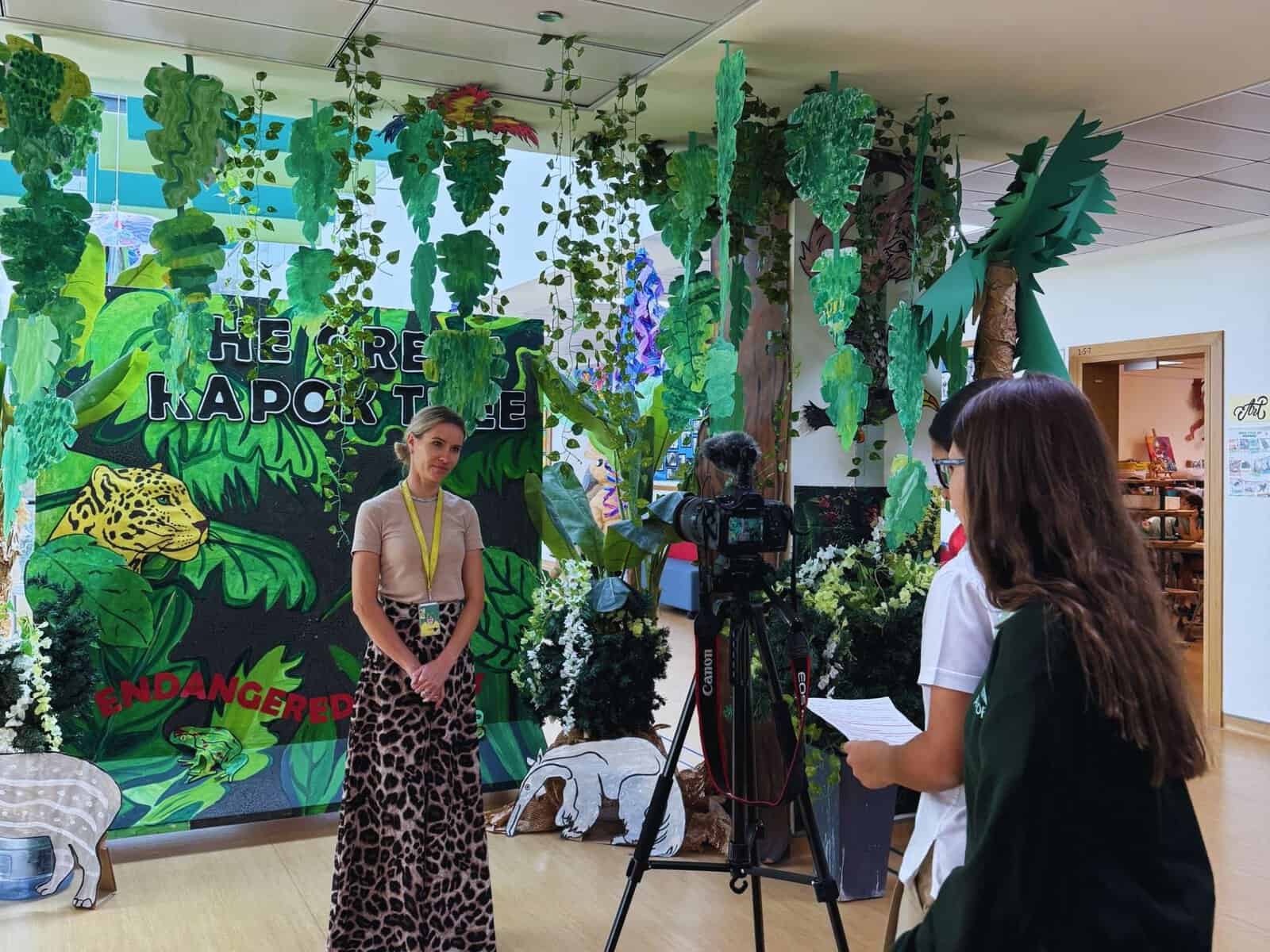 School scene with jungle-themed backdrop and students filming.