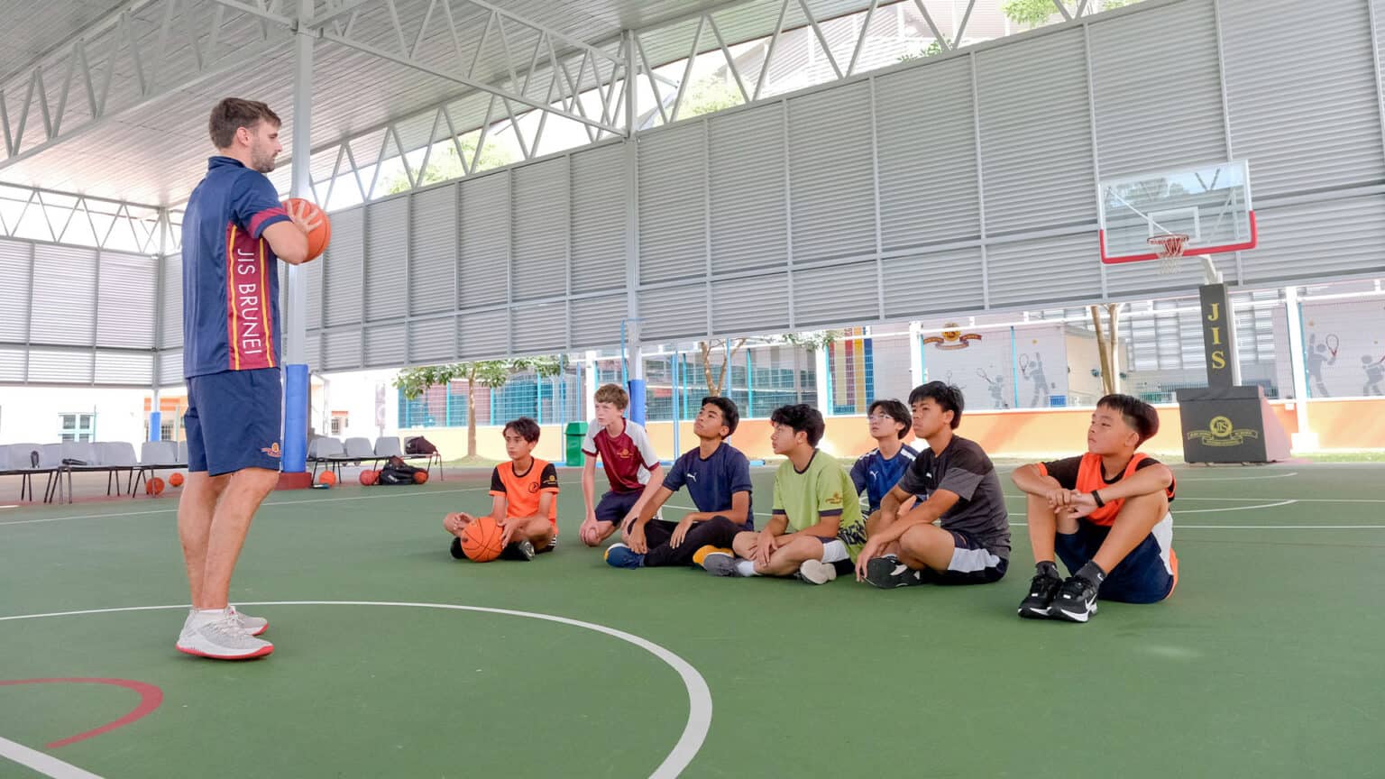 Bright indoor basketball court with young students seated on the floor, listening to coach. School sports facilities promote active learning.