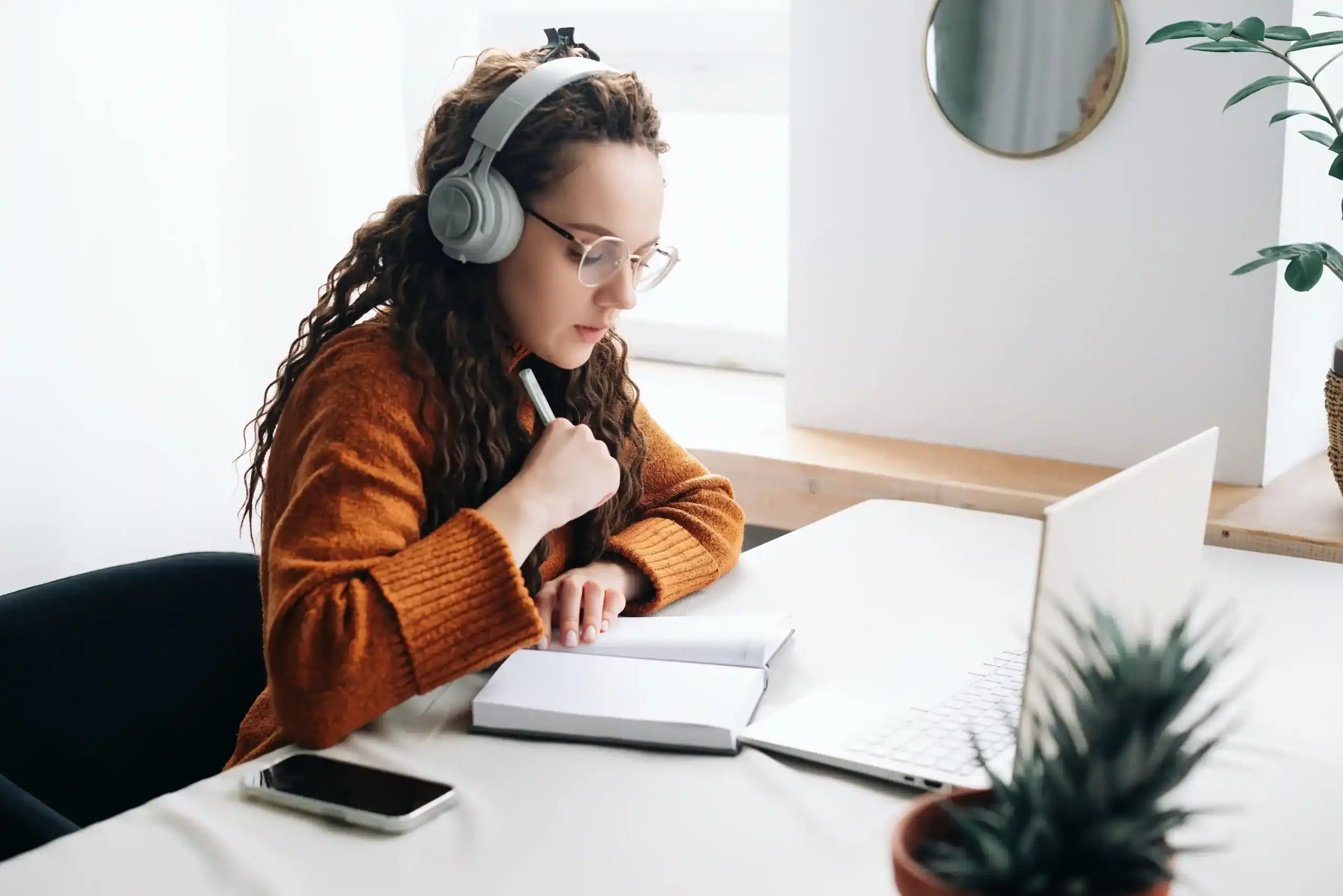 Woman studying with headphones, open laptop, notebook, and phone on white table at home, modern learning environment.