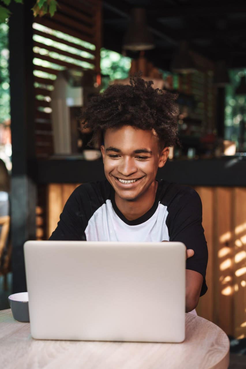 Youthful man smiling while working on a laptop in a cozy cafe setting.