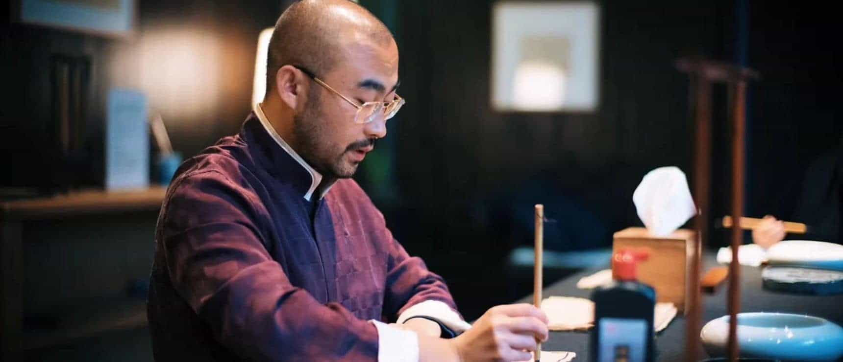 Serious man in traditional Asian attire practicing calligraphy at a modern study table.