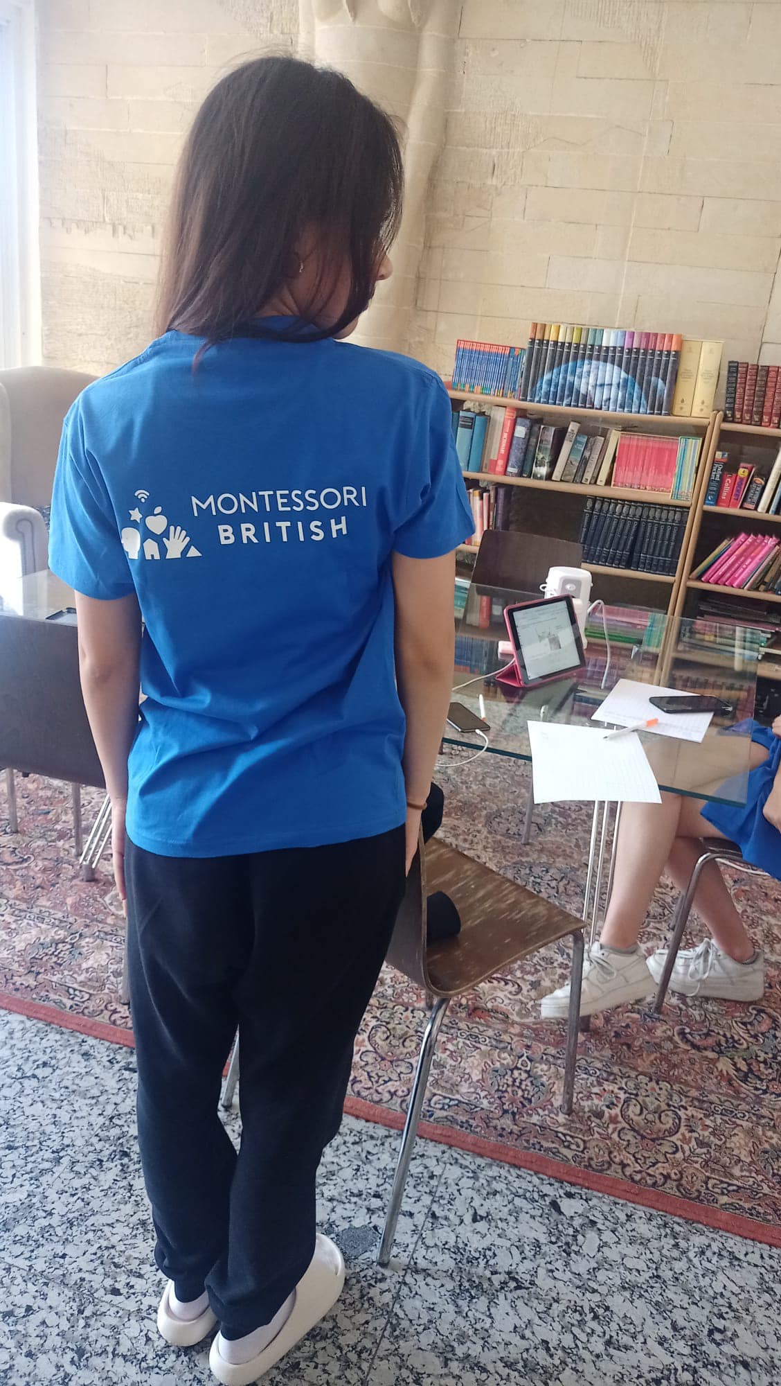 Student in Montessori British uniform in classroom with books and tablet.