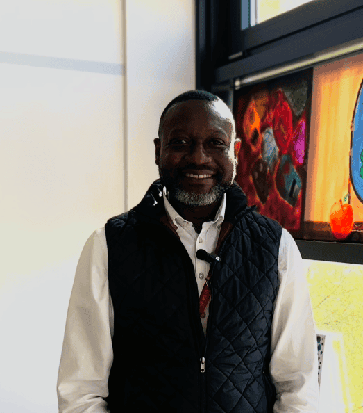 Smiling man in a classroom with colorful artwork and large windows.