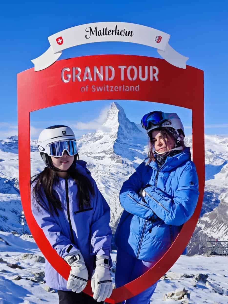 Switzerland Matterhorn mountain with two girls in ski gear inside a red "Grand Tour of Switzerland" frame.