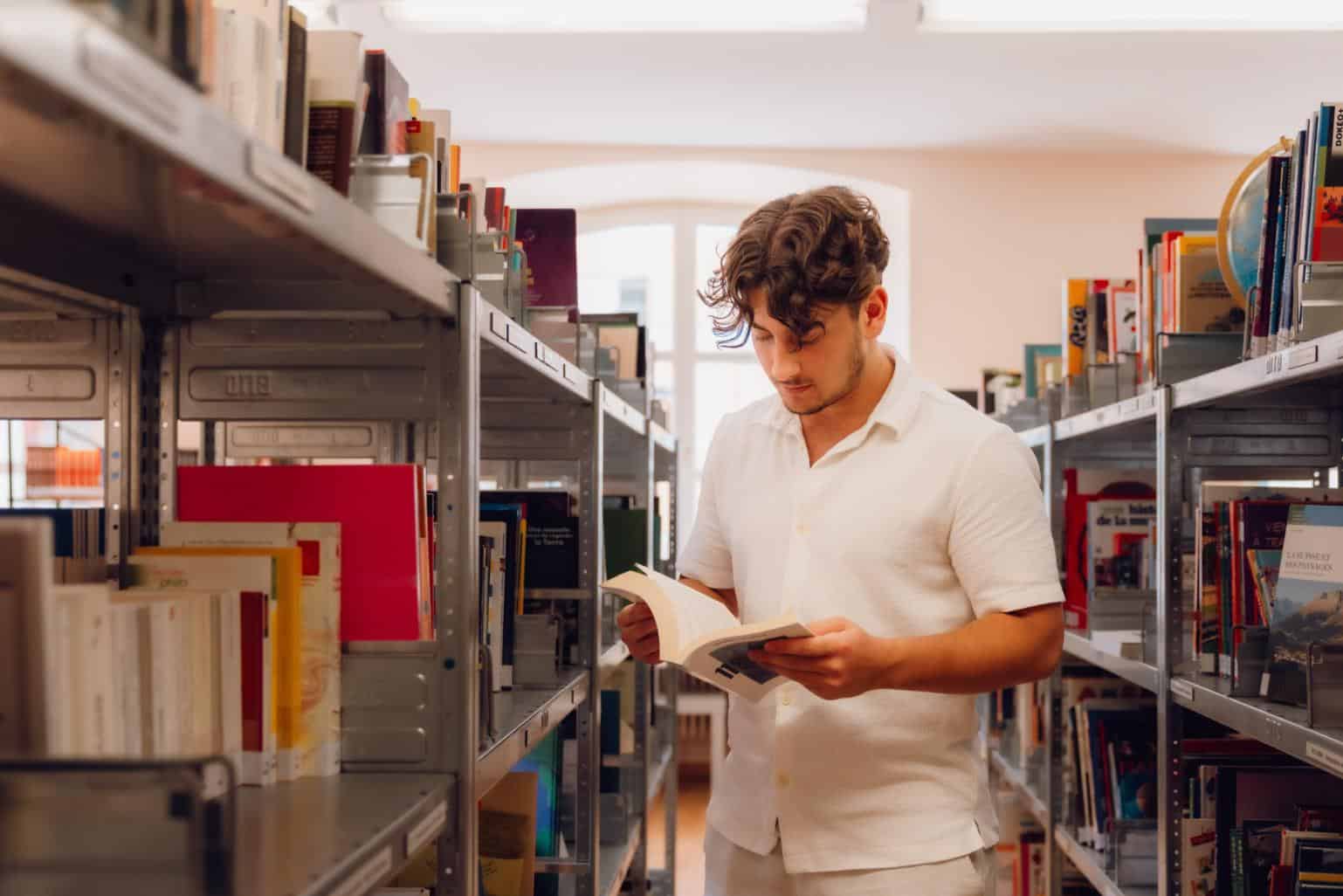 Young man reading a book in a library with shelves filled with books, promoting world school learning and education.