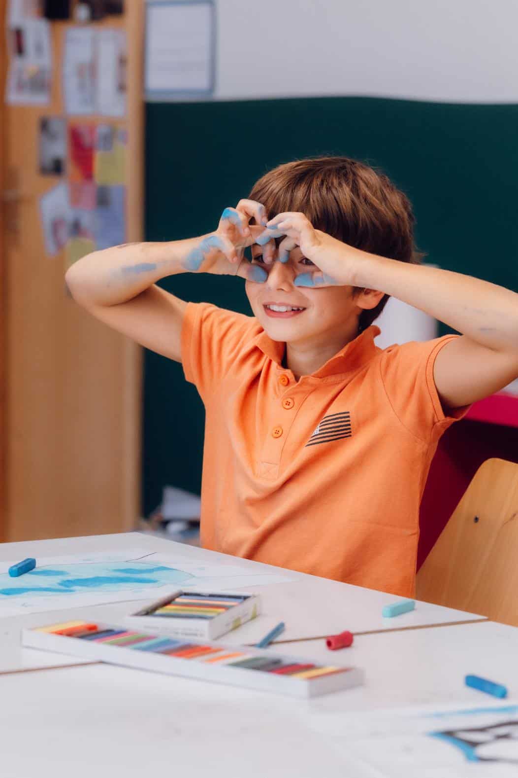 Colorful schoolboy making a playful binoculars gesture during art class.
