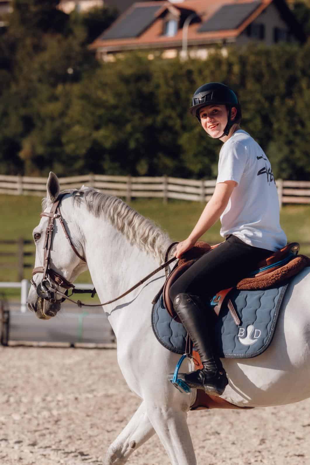Happy young girl riding a white horse outdoors at a school riding center, wearing a helmet and casual clothes.