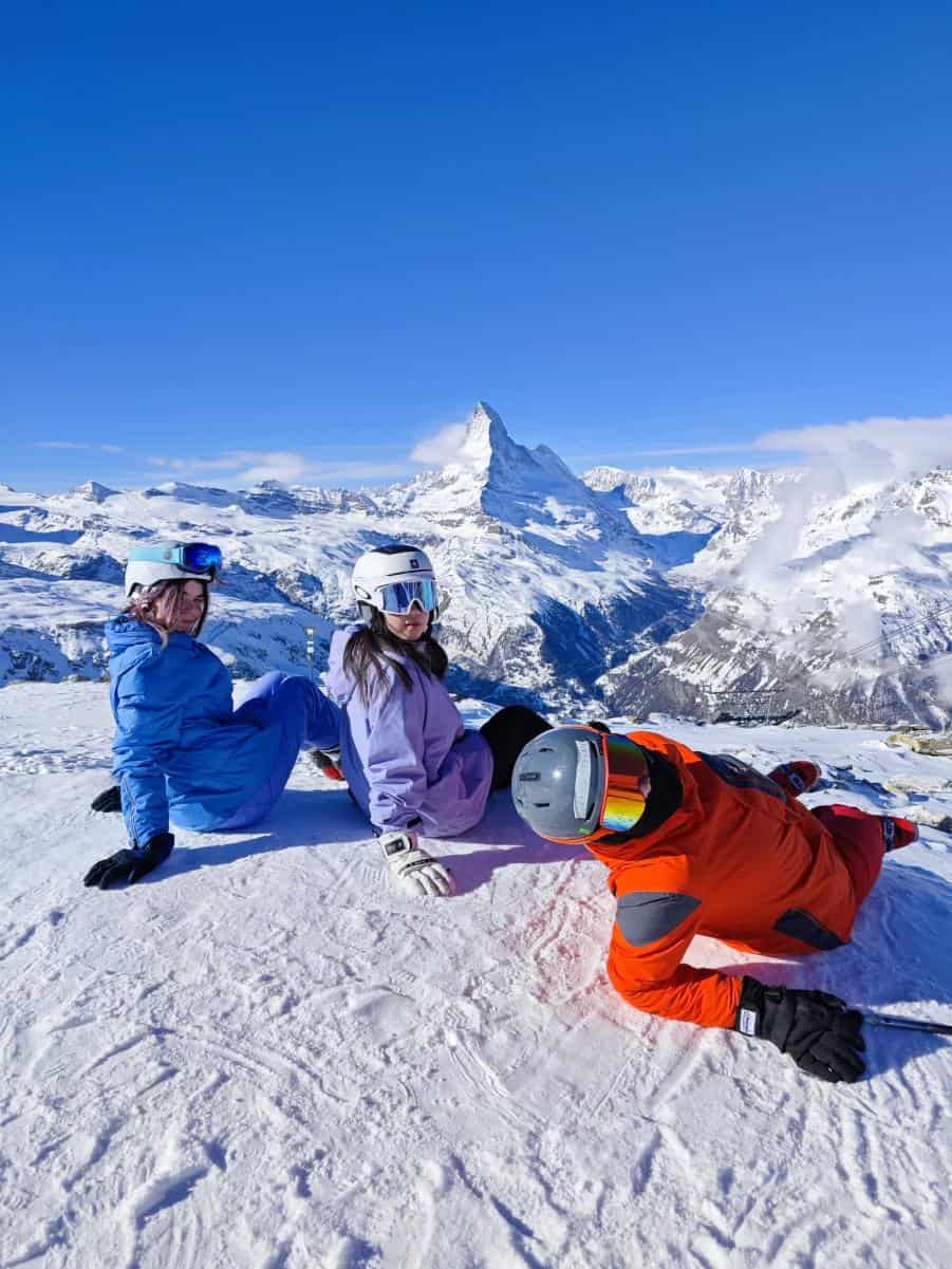 Kids in colorful ski gear lying on snow with snowy mountains and Matterhorn in background, exploring winter sports.