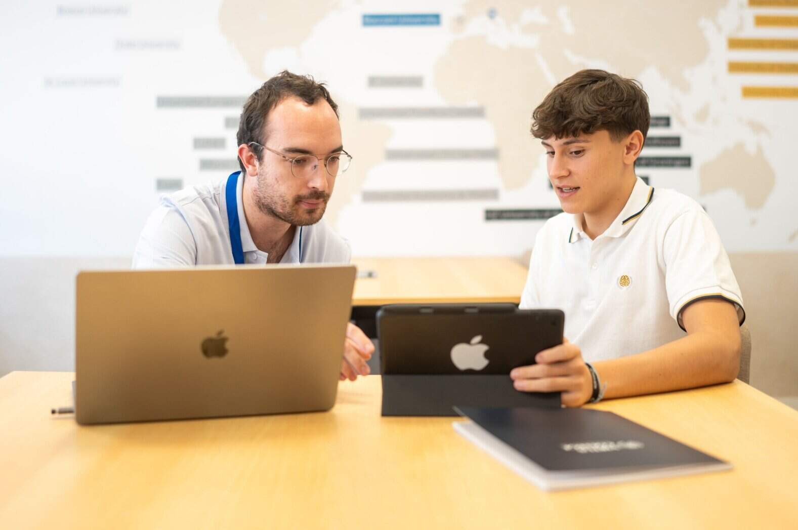 Two students working together with laptops and tablet in a classroom setting.